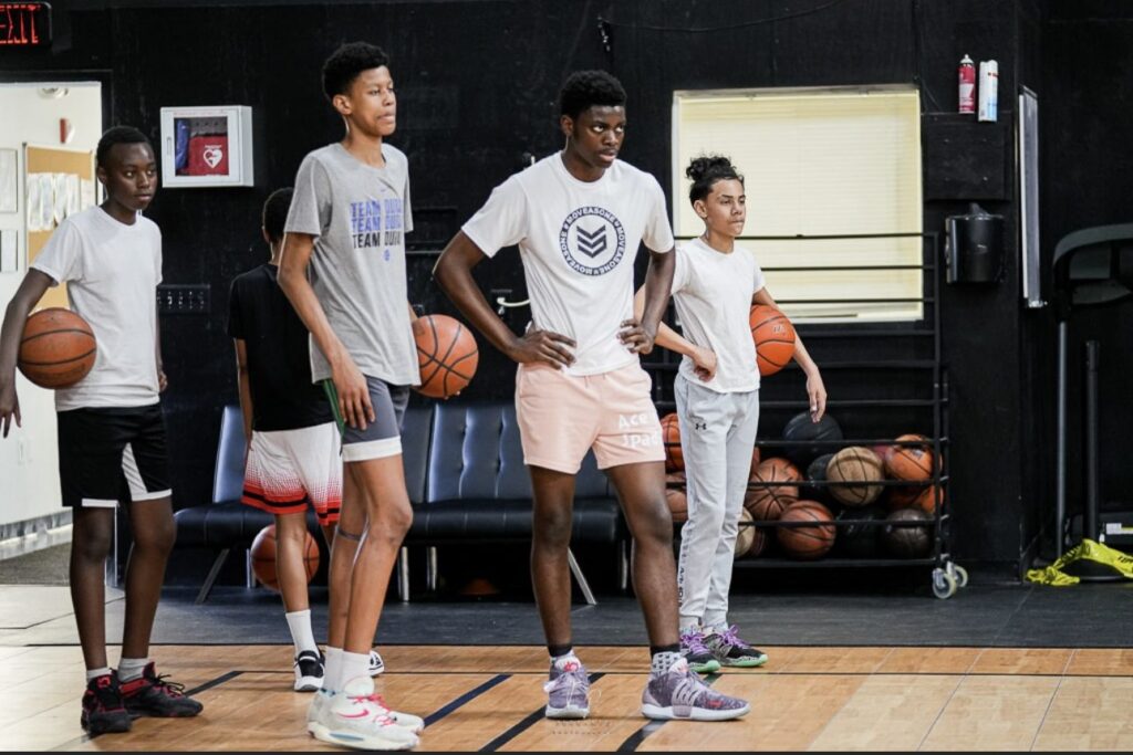 Young players lined up with basketballs in an indoor training facility, wearing team-branded shirts.