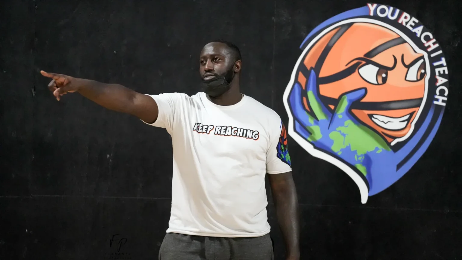Coach wearing a "Keep Reaching" shirt pointing, with the You Reach I Teach logo on the wall.